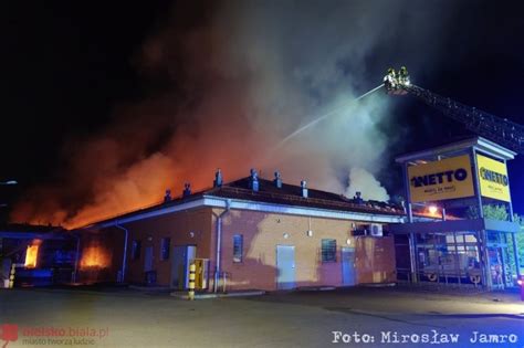 widok na spalony sklep Netto z zawalonym dachem, widoczne wozy strażackie i dym nad ulicą Piaskową