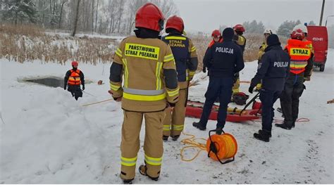 Grupa strażaków nurkujących pod lodem podczas szkolenia, z widocznym wyciętym przeręblem i asekuracją