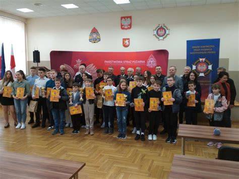 Zdjęcie z Turnieju Wiedzy Pożarniczej w Sokolnikach, pokazujące uczniów, dyplomy, puchary oraz Andrzeja Waligórę wręczającego nagrody