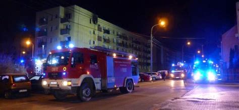 Zdjęcie przedstawiające akcję gaśniczą strażaków w nocy przy budynku mieszkalnym, z widocznym dymem i światłami służb ratunkowych.
