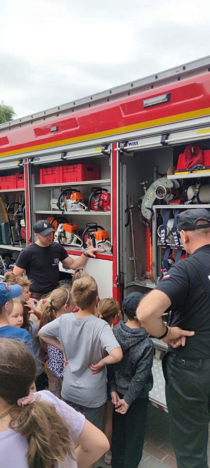 Zdjęcie dzieci zwiedzających remizę strażacką, oglądających wozy strażackie i sprzęt.