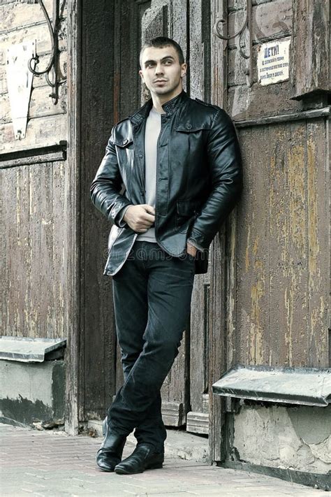 Młody mężczyzna w skórzanej kurtce typu biker, ciemnych jeansach i czarnym t-shircie, stojący na tle neonowego oświetlenia klubu.