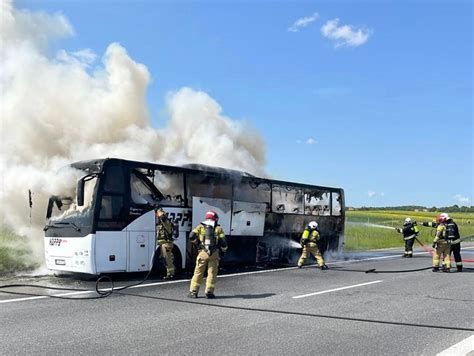 Zdjęcie autokaru objętego pożarem na autostradzie, z widocznymi strażakami i pianą gaśniczą