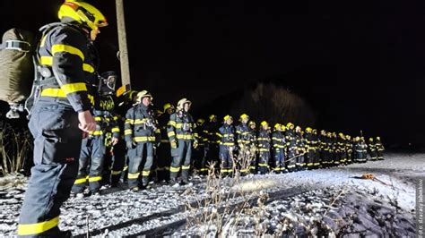 Strażacy w pełnym rynsztunku gaśniczym, wyposażeni w aparaty oddechowe, podchodzący do płonącego samochodu, z dymem i płomieniami