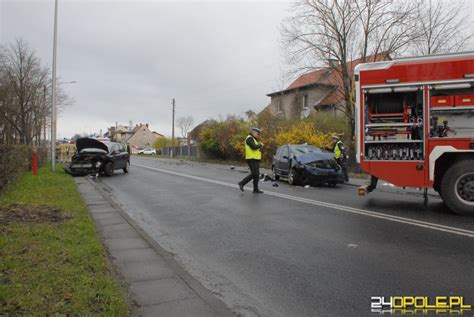 Zasłonięty widok na miejsce wypadku drogowego z udziałem dwóch samochodów osobowych, z włączonymi światłami awaryjnymi pojazdów służb ratunkowych.