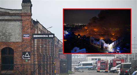 The historical hall of former Railway Rolling Stock Repair Works in Gdańsk Przeróbka engulfed in flames, with thick smoke rising. Firefighters operating from the ground and water.