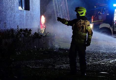 Strażacy OSP gaszący pożar poddasza budynku jednorodzinnego w nocy, z widocznym dymem i światłem z reflektorów