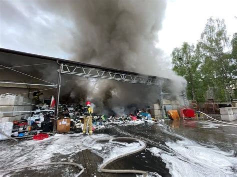 Fotografia strażaków w sprzęcie, gaszących płonące ruiny hal w Gliwicach, widoczne węże i piany, dzienny krajobraz
