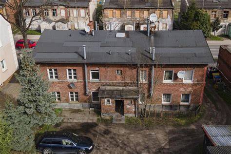 Wielopiętrowy budynek mieszkalny z widocznymi śladami pożaru w jednym z okien, otoczony wozami strażackimi i ratownikami.