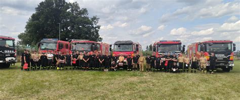 Grupa strażaków ochotników pozuje z nowymi wozami strażackimi podczas uroczystości przekazania.