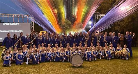 Zespołowe zdjęcie Młodzieżowej Orkiestry Dętej OSP Rajsko na tle sceny lub budynku
