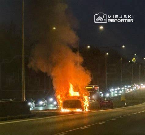 Zapalony samochód osobowy na poboczu drogi, z widocznym dymem i płomieniami