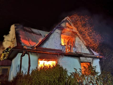Zdjęcie przedstawiające płonący budynek mieszkalny w nocy, z widocznymi strażakami i wozami strażackimi