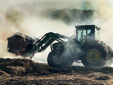 Thematic photo of a burning tractor and hay bales in a field, with firefighters in action