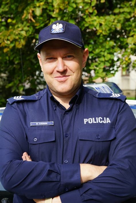 Policjant w mundurze stojący obok radiowozu, uśmiechnięty.