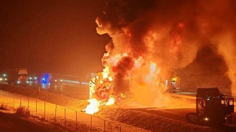 Płonąca naczepa chłodni na poboczu drogi, z widocznymi strażakami gaszącymi pożar pianą