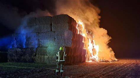 Photo showing a large stack of burning hay bales with smoke, and firefighters working