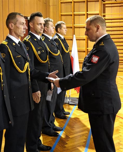 Infografika lub zdjęcie z ceremonii wręczenia awansów strażackich, z widocznym nowym wozem bojowym w tle