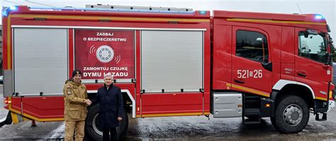 Zdjęcie przedstawiające współczesny wóz strażacki zaparkowany przed remizą